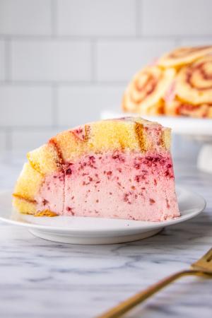 A side view of a slice of Raspberry Charlotte cake, featuring pink Bavarian cream under a layer of vanilla chiffon swiss cake roll, sits on a white plate on a marble counter with a gold fork in the foreground. In the background, the remainder of the cake on a white cake stand can be seen just in front of a white subway tiled backsplash.  