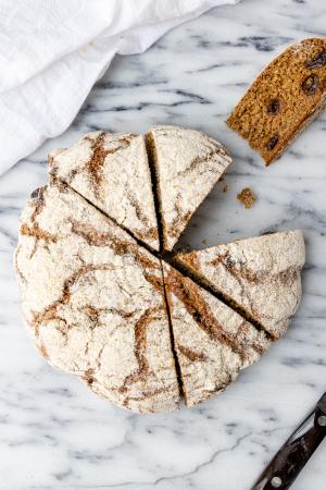 A round loaf of whole grain raisin bread partially cut into wedges with one wedge on its side to reveal its crumb